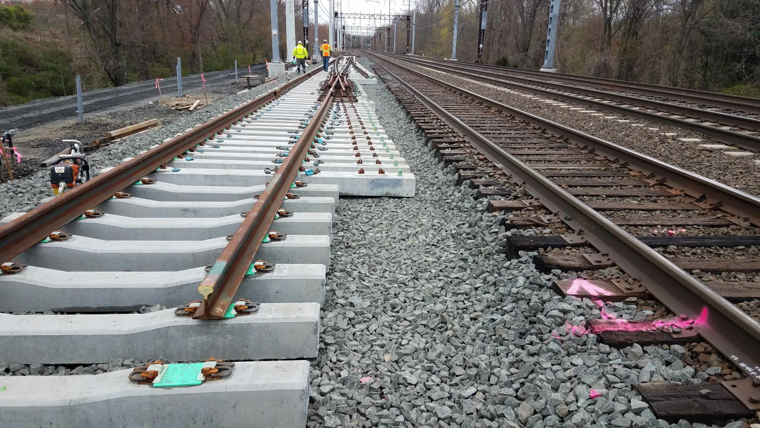 Construction site image of a the main line. In the foreground are new rails and ties with electric infrastructure in the background. Additionally, two workers exist in the background.