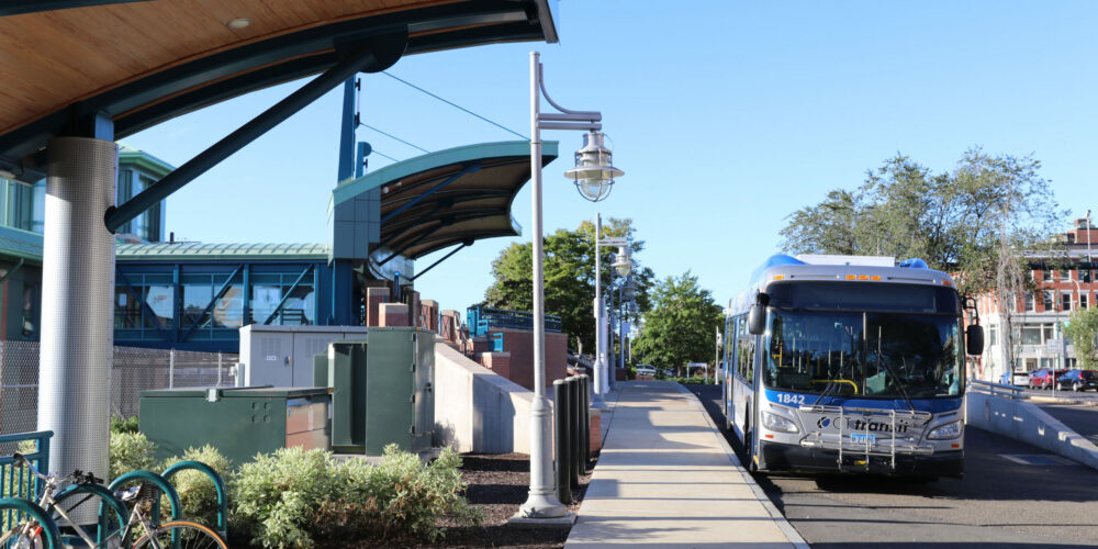A CT transit bus arrives in New Haven. The blue/grey bus is in the waiting area next to a covered bus shelter.