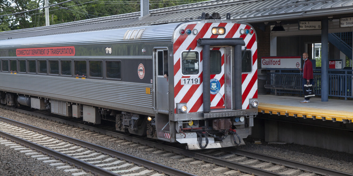 A train car sits at Guilford station. An individual waits on the platform while people board.